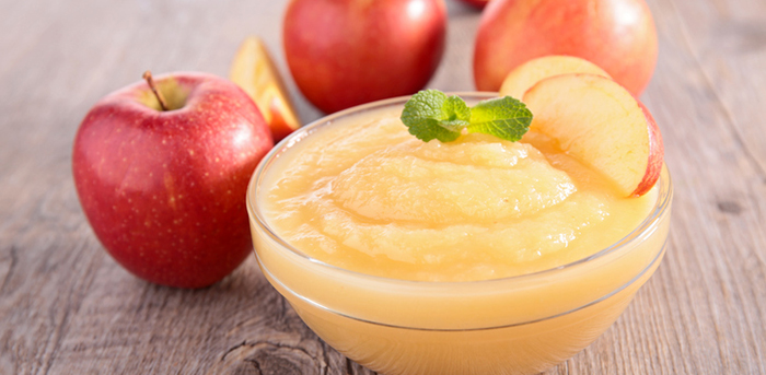 Small bowl of applesauce on wooden table surrounded by whole apples