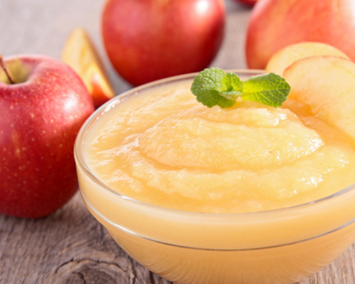 Small bowl of applesauce on wooden table surrounded by whole apples